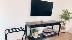 Ein Zimmer mit einem schwarzen Metallstuhl, einem Holztisch mit Ablagefläche, einer Topfpflanze und einem flachbildfernseher an der Wand.