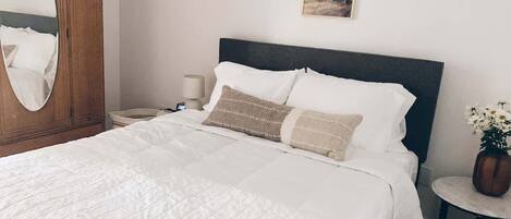 A neatly made bed with white bedding and two decorative pillows, a wooden wardrobe with a mirror, a bedside table with a lamp, and a small round table with a vase of flowers.