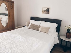 A neatly made bed with white bedding and two decorative pillows, a wooden wardrobe with a mirror, a bedside table with a lamp, and a small round table with a vase of flowers. - Beautiful Private Guest House in Arcadia (Phoenix)