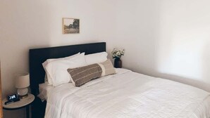 Ein ordentlich bezogenes Bett mit weißen Leinen und einem dunklen Kopfteil, ein Nachttisch mit Lampe und ein kleines gerahmtes Bild an der Wand.