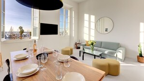 A modern living room with a dining area, a sofa, a coffee table, and a view of the cityscape. - Upstays Harbor View 83m² Apartment Palais (Cannes)