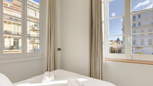A hotel room with a bed, two rolled towels, a window with a view of buildings, and curtains. - Upstays Harbor View 83m² Apartment Palais (Cannes)