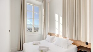 Ein Hotelzimmer mit einem großen Bett, einem Holzkopfende, einem Nachttisch und Blick auf die Stadt durch ein großes Fenster.