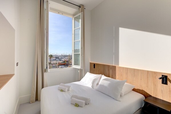 A hotel room with a large bed, a wooden headboard, a window with curtains, and a view of a cityscape.