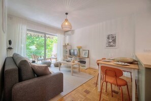 A modern living room with a sofa, a dining area with a table and chairs, a television, and a large window with a view of greenery. - Immogroom - Modern with Terrace - Air (Cannes)