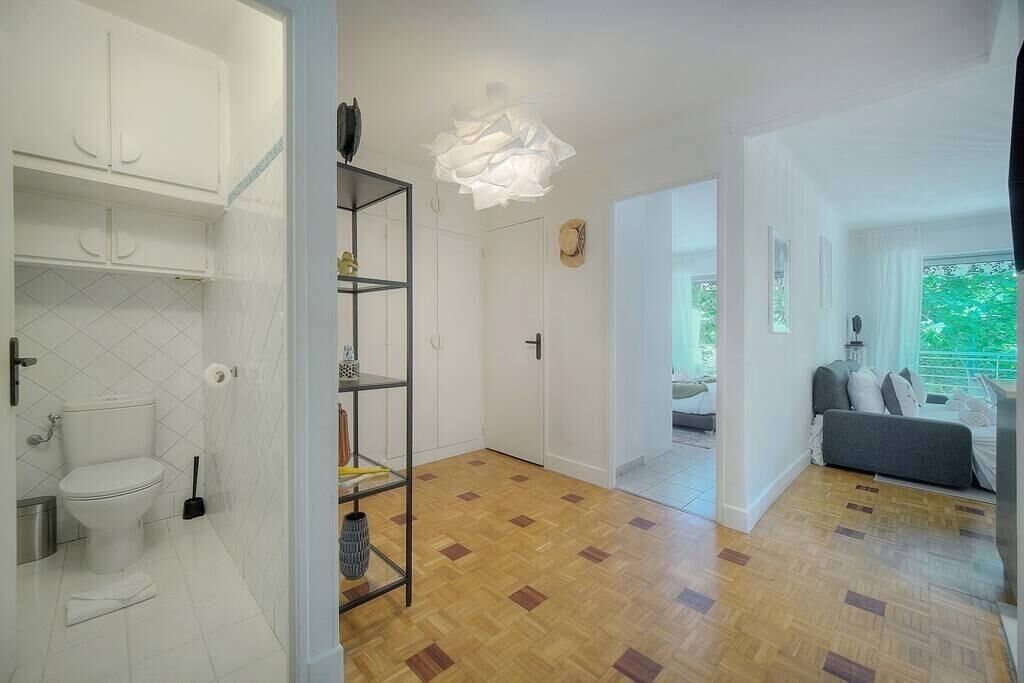 A modern interior with a wooden floor, white walls, a white toilet, a metal shelving unit, and a grey sofa in a living area with a view of greenery.