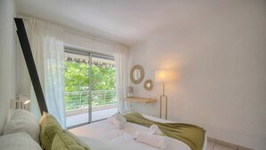Une chambre d’hôtel avec un grand lit, un bureau, une chaise, une fenêtre avec des rideaux et un balcon agrémenté de plantes.