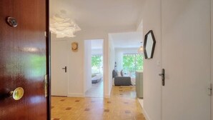 Un couloir avec une porte en bois, un mur blanc et une vue sur un espace de vie comprenant un canapé et une fenêtre.