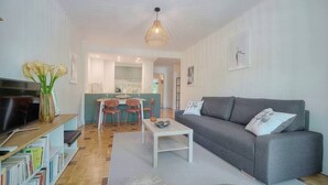 Un salon moderne avec un canapé gris, une table basse et un téléviseur posé sur une étagère en bois. On aperçoit également un coin repas avec une table et des chaises, ainsi qu’une cuisine au fond.