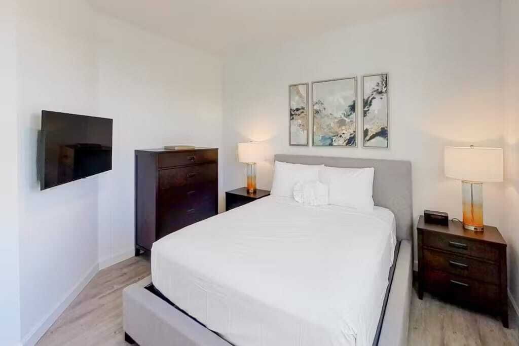 A modern hotel room with a large bed, a flat-screen TV mounted on the wall, a dark wooden dresser, and two bedside tables with lamps.