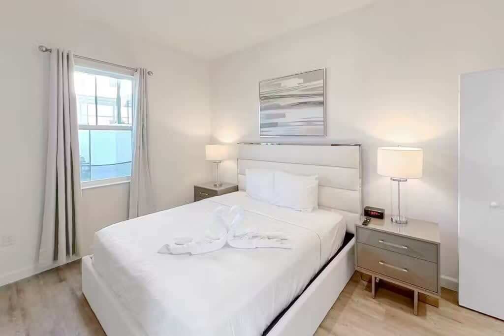 Une chambre d’hôtel comprenant un lit, deux tables de chevet avec des lampes, une petite table, une œuvre d’art accrochée au mur et une fenêtre avec des rideaux.
