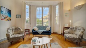 A living room with two armchairs, a sofa, and a coffee table. There are large windows with curtains, a painting on the wall, and a lamp on a side table. - Upstays Terrace Garden 100m² Villa Center (Cannes)