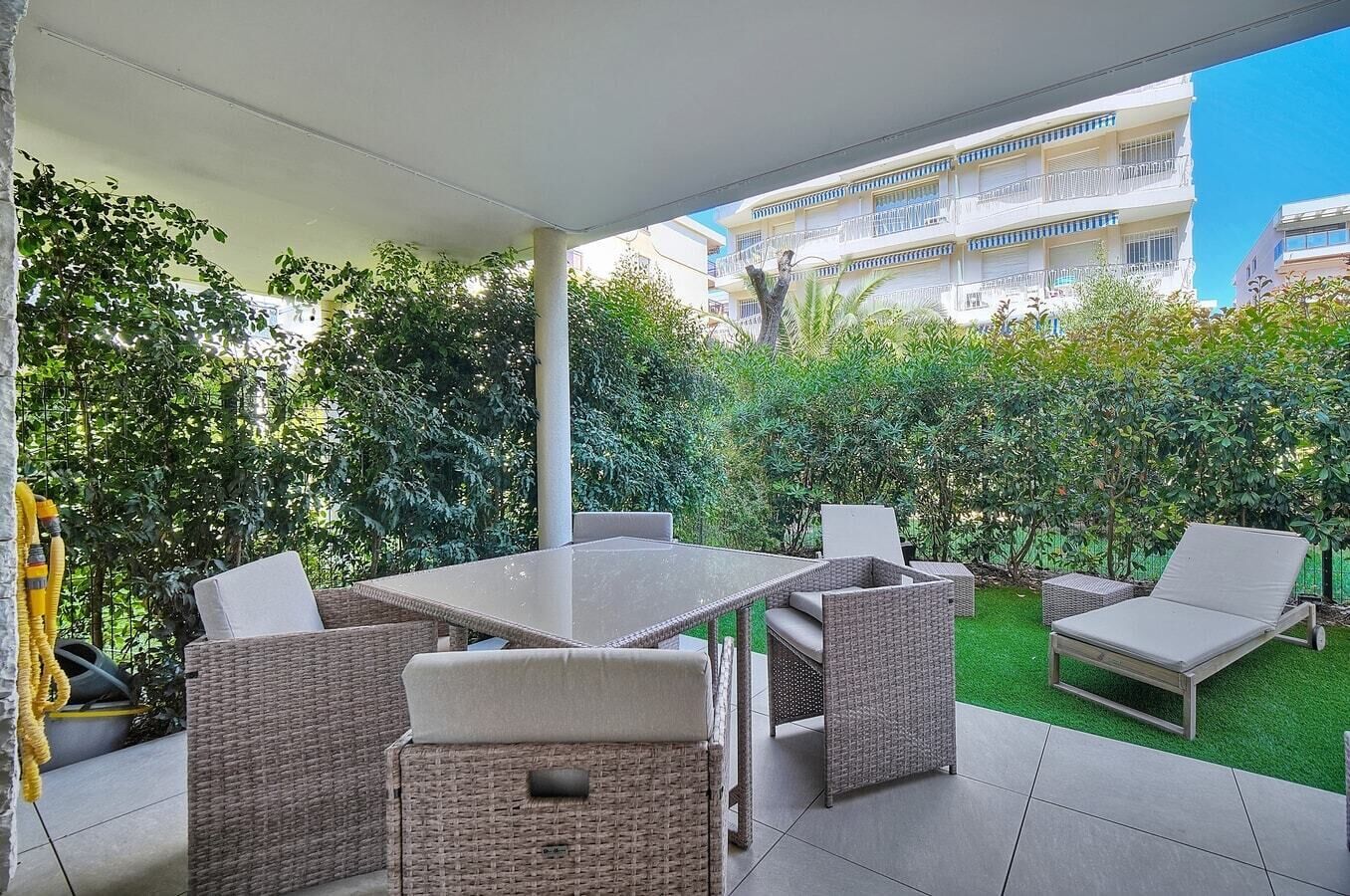 En uteplats med ett bord och stolar, omgiven av frodig grönska och med utsikt över en bostadsbyggnad.