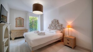 Ein Schlafzimmer mit Bett, Nachttisch, Lampe, Kommode, Spiegel und einem Fenster mit Blick auf Grünflächen.