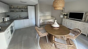 Eine moderne Küche mit weißen Schränken, ein Essbereich mit Korbstühlen und ein Flachbildfernseher auf einem weißen Schrank.