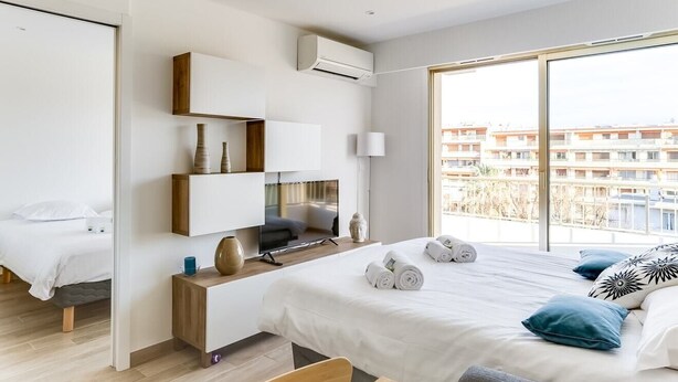 A modern hotel room with a large bed, a desk with a flat-screen TV, a wardrobe, and a view of the cityscape. - Upstays Croisette Terrace 35m² Apartment (Cannes)