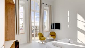 Ein modernes Hotelzimmer mit einem Bett, zwei gelben Sesseln, einem kleinen runden Tisch und einem an der Wand befestigten Fernseher.