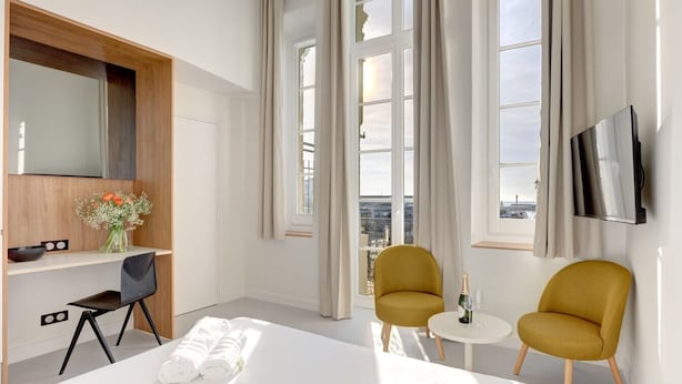 Ein modernes Hotelzimmer mit Bett, zwei Sesseln, einem kleinen Tisch, einem Fernseher und einem Fenster mit Vorhängen.