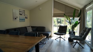 A modern living room with a wooden dining table, dark sofas, and a large window with a view of greenery. - Europarcs Kagerplassen (Warmond)
