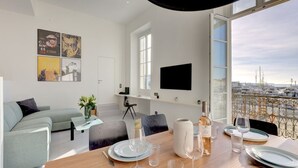 A modern living room with a dining area, a flat-screen TV mounted on the wall, and a balcony with a city view. - Upstays Harbor View 45m² Apartment Palais (Cannes)