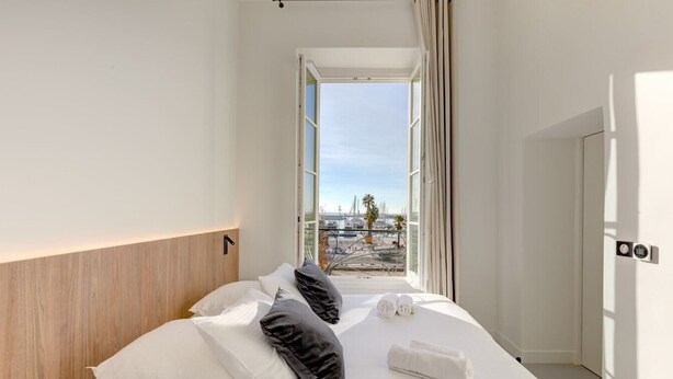 A hotel room with a bed, pillows, a towel, and a view of a cityscape through a window. - Upstays Harbor View 45m² Apartment Palais (Cannes)