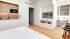 Ein modernes Hotelzimmer mit einem großen Bett, einer integrierten Küche inklusive Mikrowelle, einem an der Wand montierten Fernseher und einer kleinen Sitzecke mit Tisch und Stühlen.