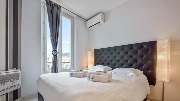 A hotel room with a large bed, a window with curtains, an air conditioning unit, and a bedside lamp.
