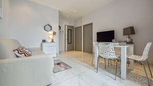 A modern living room with a white sofa, a dining table with white chairs, a flat-screen TV, and a small white cabinet.
