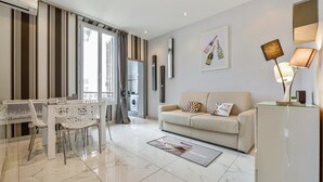 A modern living room with a sofa, a dining table with chairs, a kitchen in the background, and a large window with curtains.