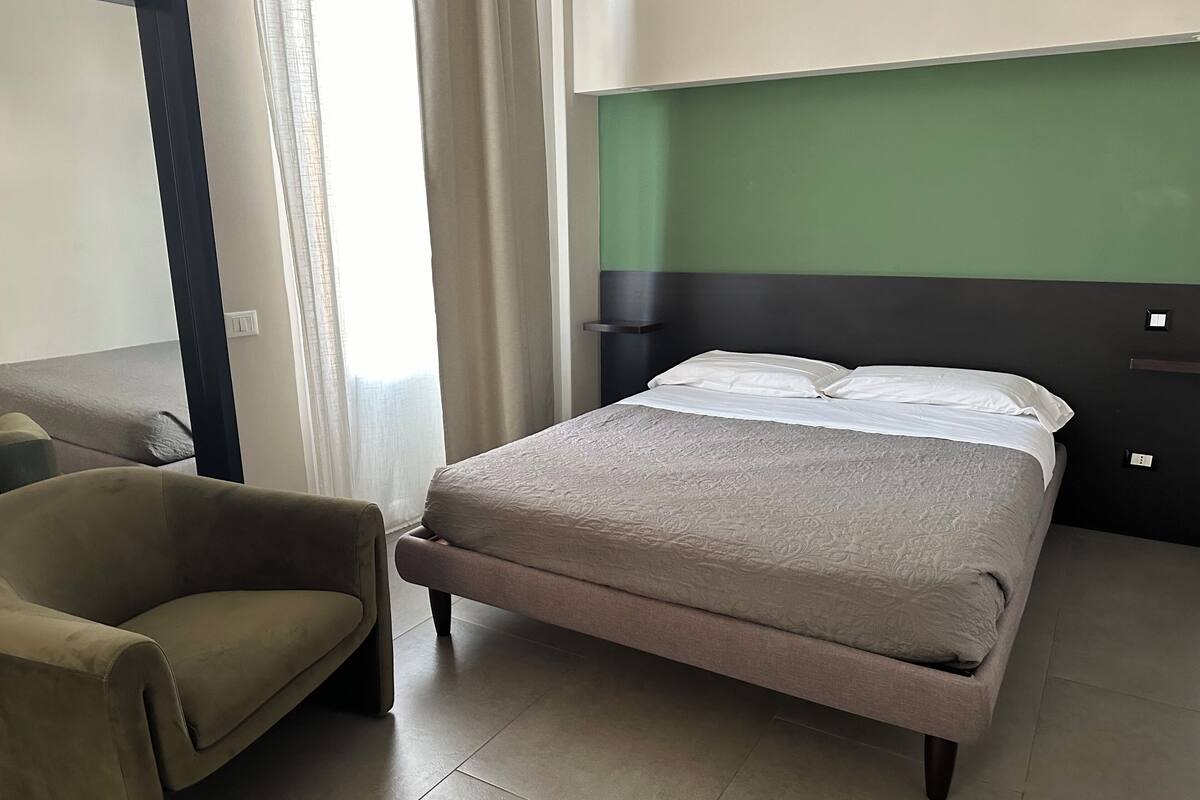 A hotel room with a bed, a chair, a green wall, and a window with curtains.