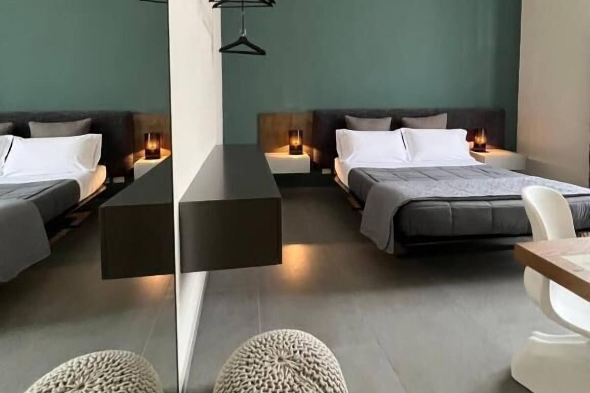 A modern hotel room with two beds, bedside tables, hanging lights, and a round ottoman.