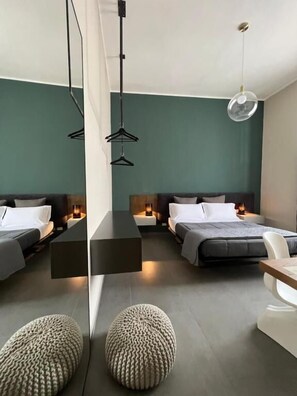 A modern hotel room with two beds, bedside tables, hanging lights, and a round ottoman. - Catania Rooms (Catania)