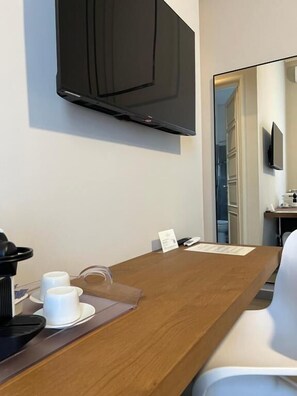 A hotel room with a wooden desk, a television mounted on the wall, a coffee machine, and a white chair. - Catania Rooms (Catania)