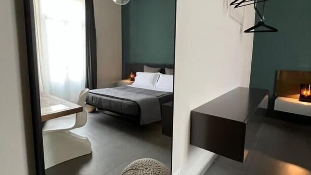 A modern hotel room with a large mirror, a bed, a bedside table, a small round ottoman, and a seating area with a bench and a table. - Catania Rooms (Catania)