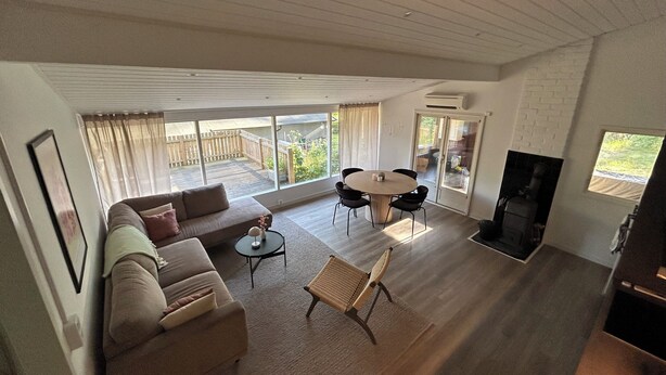 Amplia sala de estar con sofá, una mesa redonda y sillas. Cuenta con una chimenea y vistas a una terraza de madera a través de amplias ventanas.