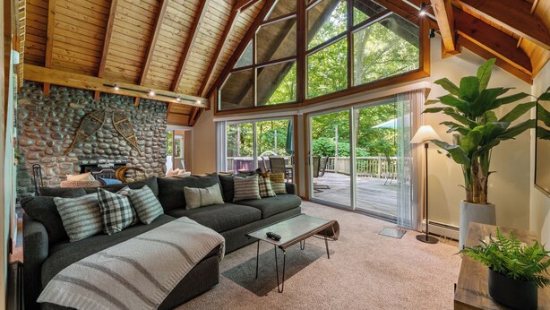 A cozy living room with a stone wall, a large window, and a wooden ceiling. - 1 Min to Camelback! Creekside Escape + FirePit & BBQ (Tannersville)