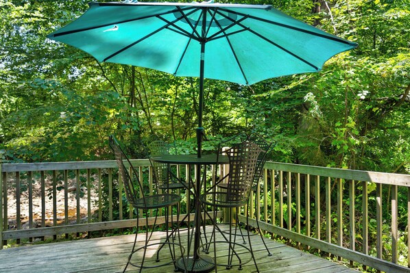 Une terrasse en bois avec une table et des chaises sous un parasol vert, entourée d’une végétation luxuriante.