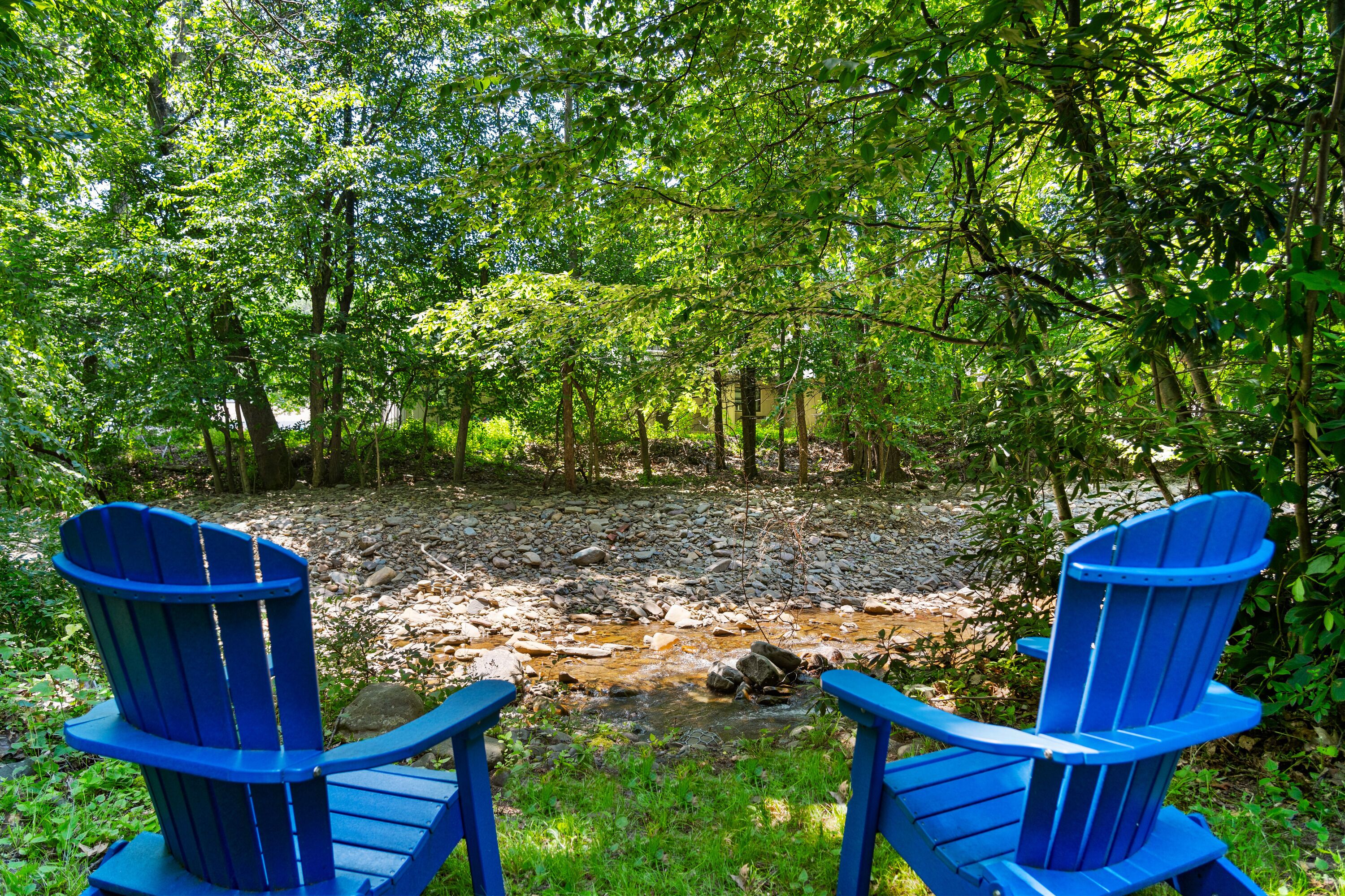 Deux chaises Adirondack bleues, placées face à un ruisseau dans une zone boisée.