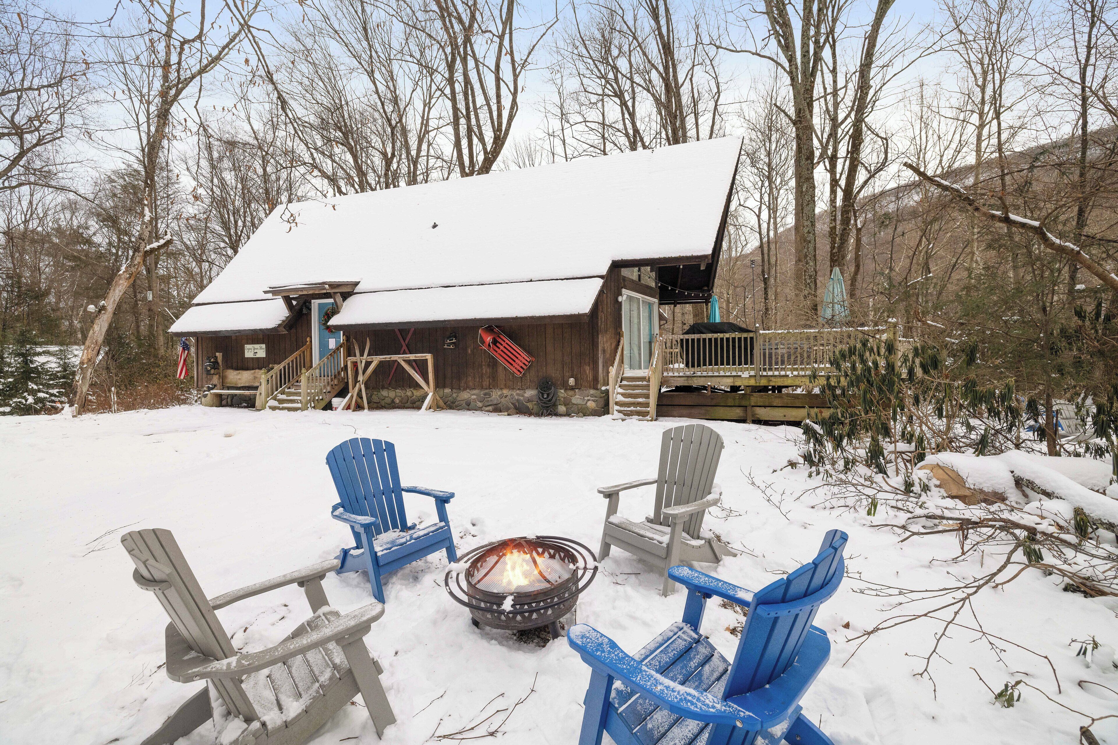 A cabin with a snow-covered roof, a fire pit in the center, and four Adirondack chairs around it.