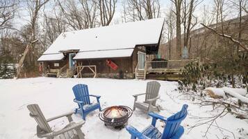 A cabin with a snow-covered roof, a fire pit in the center, and four Adirondack chairs around it.