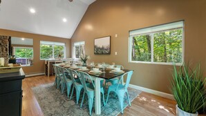 A dining area with a long table set for six, surrounded by turquoise chairs, with a large window providing a view of trees. - 1 Min to Camelback! Creekside Escape + FirePit & BBQ (Tannersville)