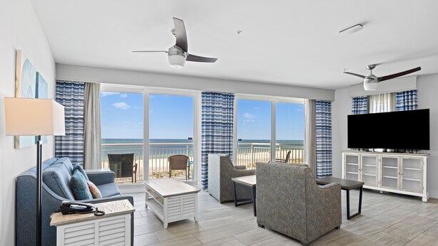A modern living room with a sofa, armchair, and TV stand, large windows with a view, and ceiling fans. - 2BR Bayview Condo with Balcony & Pool (Treasure Island)