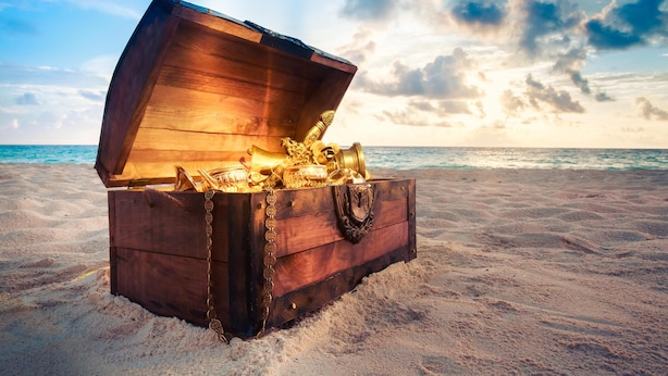 Eine hölzerne Truhe, gefüllt mit Goldmünzen, am sandigen Strand mit dem Meer im Hintergrund.