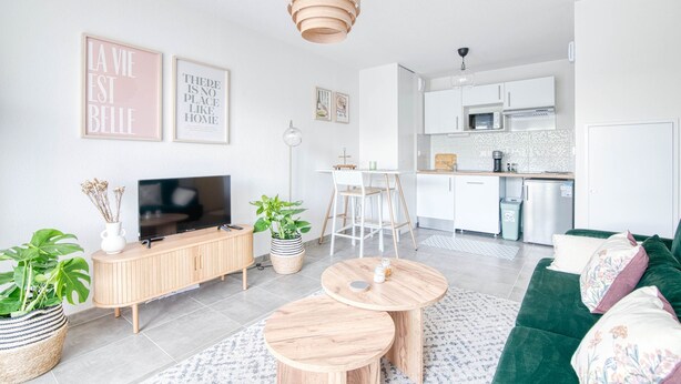 Ein modernes Wohnzimmer mit Fernseher, Holzschrank und einem grünen Sofa. Im Hintergrund ist eine Küche mit weißen Schränken und einer Mikrowelle zu sehen.