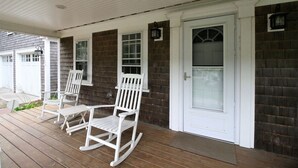 A porch with two rocking chairs, a white door with a glass panel, and a brick wall.