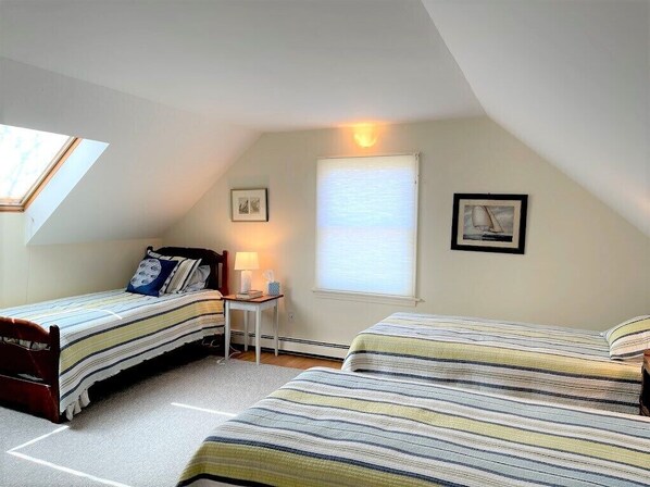 A bedroom with two beds, a skylight, a window with blinds, and a small table with a lamp. - Beautiful Cape Cod Summer Home (Orleans)