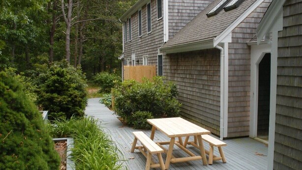 A wooden deck with a picnic table and benches, surrounded by greenery and a house with a shingled roof.