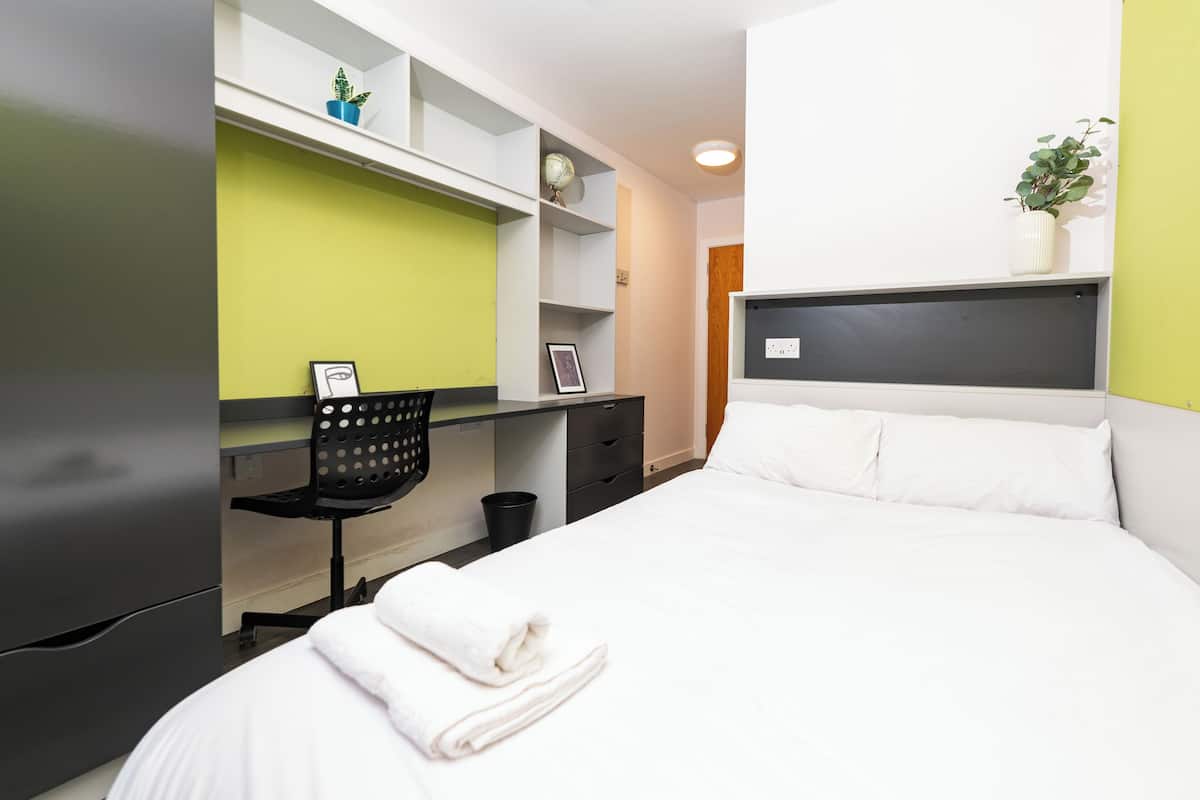 Une chambre d’hôtel comprenant un lit, un bureau avec une chaise, une petite étagère avec des plantes et une table de chevet.