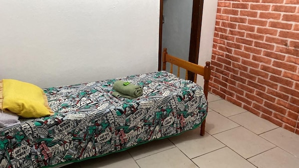 A small room with a bed, a wooden chair, a yellow pillow, and a brick wall.