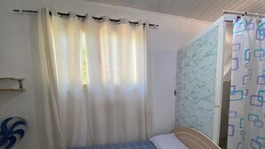 Een eenpersoonsbed met een blauwe bedspreide met ruitpatroon, twee opgerolde gele handdoeken en een wit kussen. Een wit gordijn bedekt gedeeltelijk een raam, en rechts is een gordijn met dessin zichtbaar.